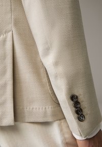 Manica di un blazer beige con quattro bottoni scuri, tessuto texturizzato, indossato sopra pantaloni beige abbinati.