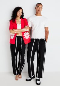 Camisa corta de manga corta con bordado floral rojo, top corto blanco, pantalones negros con rayas onduladas blancas y calzado en contraste.