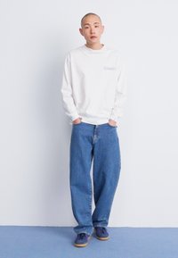 Fehér, hosszú ujjú ing, kis grafikával, párosítva laza kék farmernadrággal és sötét edzőcipővel, amelyek gumi talpúak. Egyszerű dizájn.