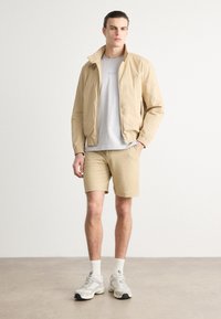 Chaqueta de cremallera color beis claro, camiseta gris, pantalones cortos beige texturizados y zapatillas ligeras. Diseño minimalista con puños elásticos y una marca sutil.