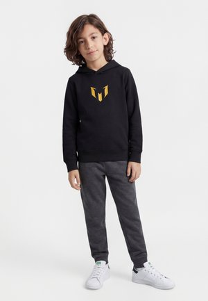 Barn med mellembrunt hår i skulderlængde iført en sort hættetrøje med gult emblem, mørkegrå bukser og hvide sneakers, stående mod en hvid baggrund.