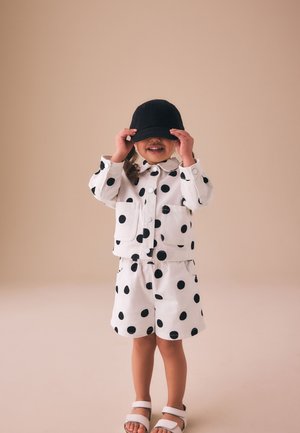 Barn iført et hvidt outfit med store sorte polkaprikker, holder en sort kasket ned over ansigtet og smiler, står på en ensfarvet beige baggrund.