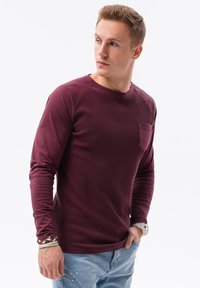 Bordeaux langekous T-shirt met ribbelstructuur, ronde hals en een kleine borstzak. Gecombineerd met lichtblauwe jeans en polsaccessoires.