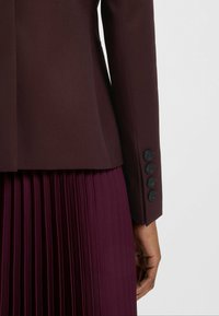 Burgundowy żakiet o dopasowanym kroju, teksturowany materiał, z czterema czarnymi guzikami na mankietach, w zestawie z pasującą plisowaną spódnicą.