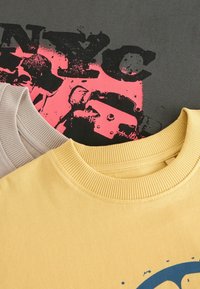 Kolme crewneck-t-paitaa eri väreissä: keltainen, beige ja harmaa. Harmaassa t-paidassa on vaaleanpunaiset graafiset tekstit ja roiskeet.