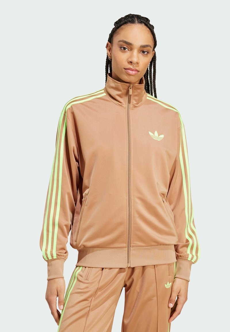 Veste de survêtement marron en matériau lisse avec des rayures vertes le long des manches. Présente un col haut et une fermeture éclair à l'avant. Logo Adidas affiché.
