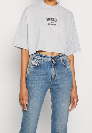 Personne portant un t-shirt court gris clair avec le texte « San Diego 1995 California », associé à un jean bleu taille haute et un tatouage visible sur le bras gauche.