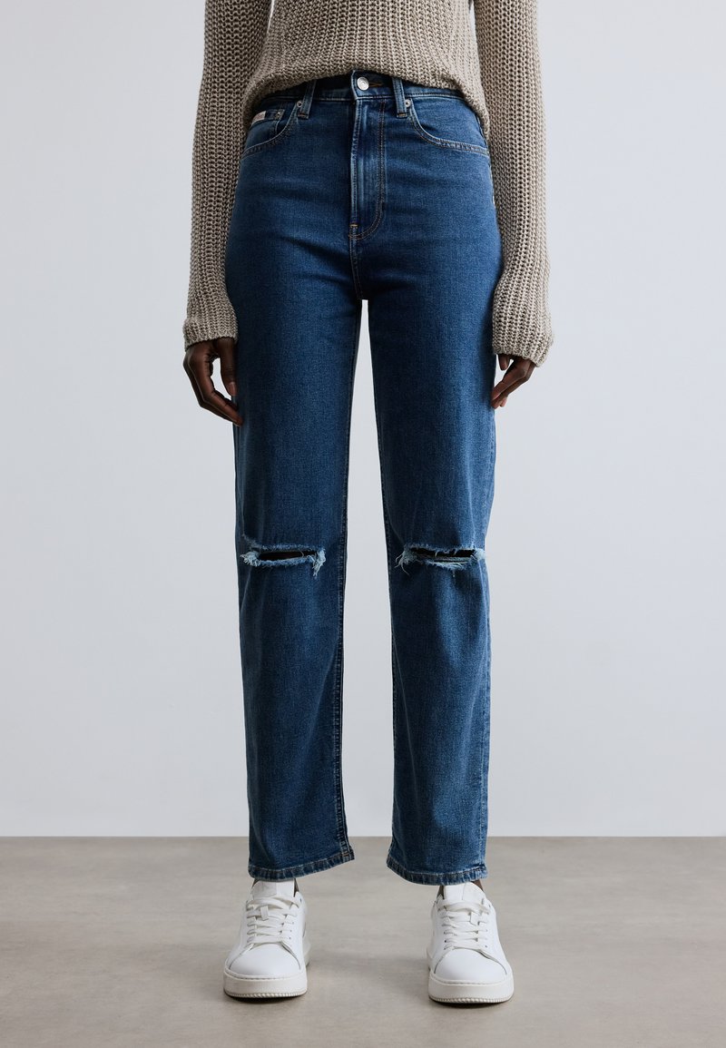 Kék farmernadrág magas derékkal, kopott térdrésszel. Fehér tornacipővel, semleges háttéren.