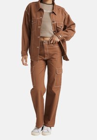 Giacca in denim marrone con tasche, abbinata a pantaloni a vita alta dello stesso colore con tasche laterali e cuciture bianche. Indossata sopra una maglietta chiara.