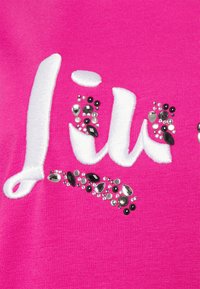 Jasně růžová látka s bílým vyšitým textem a ozdobnými broušenými kameny v černých, průhledných a stříbrných tvarech, které dodávají texturu a vizuální zajímavost.