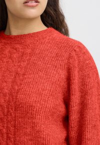 Pull en maille rouge avec un col rond, présentant des motifs texturés et une finition douce et duveteuse. Le tissu semble chaud et confortable.