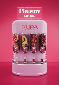 Distributore di olio per labbra con esterno rosa, quattro contenitori trasparenti pieni di frutta e piccole bottiglie chiare sotto etichettate "Pupa".