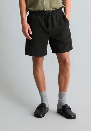 Man wearing black textured shorts, olive green shirt tucked in, gray crew socks, black slip-on shoes, standing against a plain light gray background.