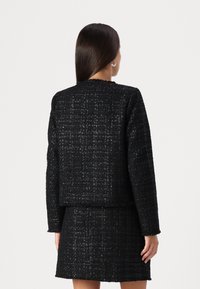 Mustat tweed-takit, joissa on laatikkomainen muotoilu, teksturoitu kangas ja suora helma, yhdistettynä matching-hameeseen, nähtynä takaa.