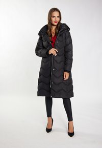 Kvinde iført lang sort quilted frakke over rød top og sorte bukser, stående med den ene hånd nær lynlåsen, på hvid baggrund.