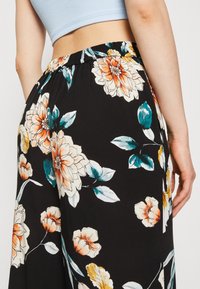 Pantalons noirs à fleurs avec des fleurs orange vives et crème, accompagnées de feuilles vertes. Taille élastique et tissu léger pour plus de confort.