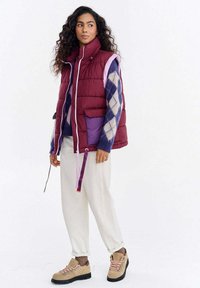 Kamizelka pikowana w kolorze bordowym z fioletowymi akcentami i białym zamkiem, noszona na wielokolorowym swetrze w wzór argyle. Połączona z kremowymi spodniami i beżowymi butami.