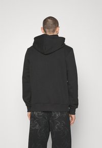 Felpa nera con un ampio cappuccio regolabile, polsini e orlo a coste, realizzata in morbido tessuto. Indossata con jeans scuri a fantasia.