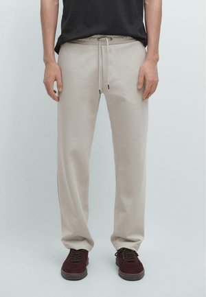 Homme portant un pantalon beige à cordon, un t-shirt noir et des baskets rouge foncé, debout sur un fond clair uni.