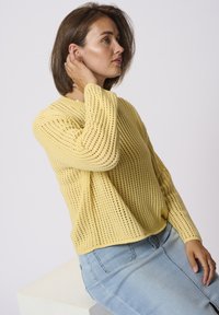 Suéter amarillo de punto con un diseño suelto y de calado. Mangas largas y escote redondeado, combinado con denim azul claro. Pose sentada.