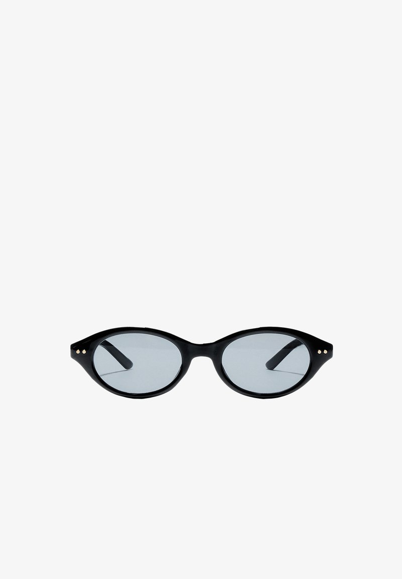 Lunettes de soleil ovales noires avec montures fines et petits points dorés près des charnières, avec verres légèrement teintés.