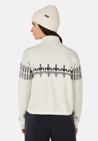 Cream sweater with black geometric pattern along shoulders, high collar, and ribbed cuffs, paired with a cream knit beanie.