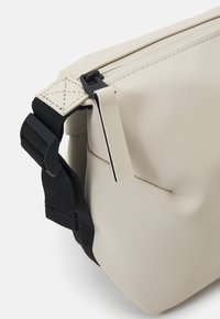 Borsa impermeabile beige con superficie testurizzata, chiusura zip e tracolla nera regolabile. Presenta un design elegante e minimalista senza alcun marchio visibile.