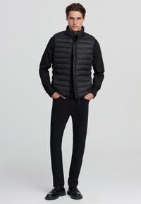 Gilet imbottito nero con collo alto, design trapuntato e chiusura con zip. Indossato sopra una maglia nera a maniche lunghe abbinata a pantaloni e scarpe nere.