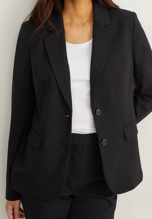Kvinde iført en skræddersyet sort blazer med to knapper over en hvid top, kombineret med matchende sorte bukser, står mod en neutral baggrund.