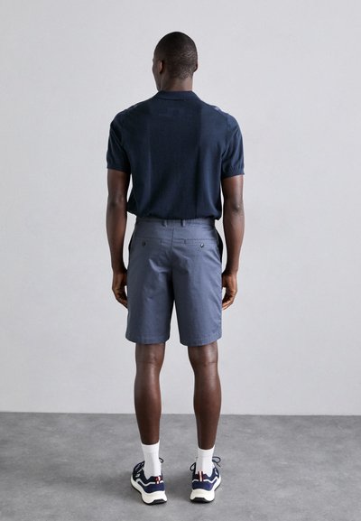 Marineblaues Polohemd mit kurzen Ärmeln, kombiniert mit grauen Shorts mit Gesäßtaschen. Das Modell trägt weiße Socken und Sneakers mit dunkelblauen Akzenten.