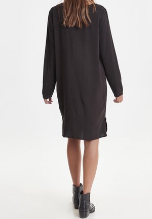 Mujer con un vestido negro de manga larga y holgado y botas negras hasta el tobillo, de pie de espaldas al espectador.