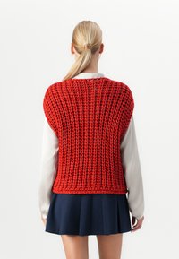Žena s blond copom oblečená v červenej hrubej pletenej veste cez bielu dlhú tričko a námornícku plisovanú sukňu, videná zozadu.