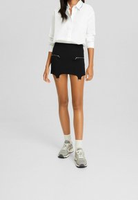 Minigonna nera con cerniere argentate su entrambi i lati, abbinata a una camicia bianca con colletto e sneakers grigie, che mette in mostra un look casual e stratificato.