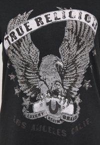 Svart t-shirt med en stor örn grafik prydd med rhinestones, texten "TRUE RELIGION" och stjärnor. Textbannerna läser "LUXURY DENIM BRAND LOS ANGELES CALIF."