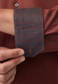 Poignet de chemise de couleur rouille texturé, présentant un tissu contrastant bleu marine avec un motif répété de dessins en tourbillon rouge. Détail du bouton visible à l'avant.