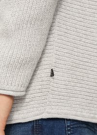 Grauer Strickpullover mit gerippter Textur, versehen mit einem schwarzen Logotag an der Seite und einem leicht abgerundeten Saumdetail.