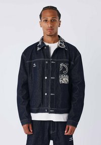 Donkerdenimjack met een contrasterende stikseldesign, voorzien van een paisleypatroon en drukknoopjes. Witte t-shirt met ronde hals eronder.