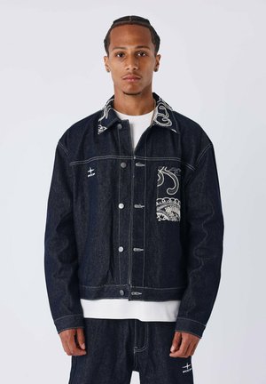 Chaqueta de mezclilla oscura con un diseño de costuras en contraste, que presenta un patrón paisley y botones a presión. Camiseta blanca de cuello redondo debajo.