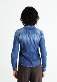 Camicia di jeans con un design fitted, colore blu medio, maniche lunghe, polsini con bottoni e una leggera sfumatura sulla parte posteriore.