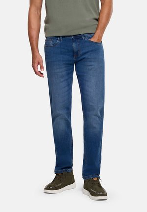 Hombre vestido con jeans ajustados azules, zapatos verde oliva con suelas blancas y una camisa verde oliva, de pie con una mano en el bolsillo.