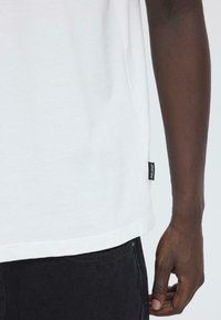 Witte katoenen T-shirt met een ronde hals, korte mouwen en een zwart label aan de zijkant. Textuurstof met een fijne afwerking.
