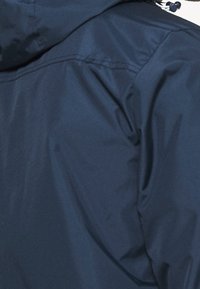 Veste imperméable navy avec une texture lisse, capuche ajustable et détails de couture visibles. Comprend une fermeture éclair et un cordon de serrage.