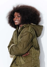 Veste matelassée olive verte avec motif coussiné, dotée d'une capuche et d'une doublure en fausse fourrure. Devant boutonné et manches longues avec poignets texturés.