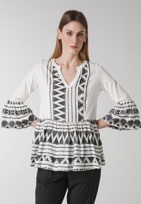 Vit blus med svart tribalbroderi, med V-ringning, utsvängda ärmar och en draperad midja. Mjuk, vävd tyg med en strukturerad mönster.