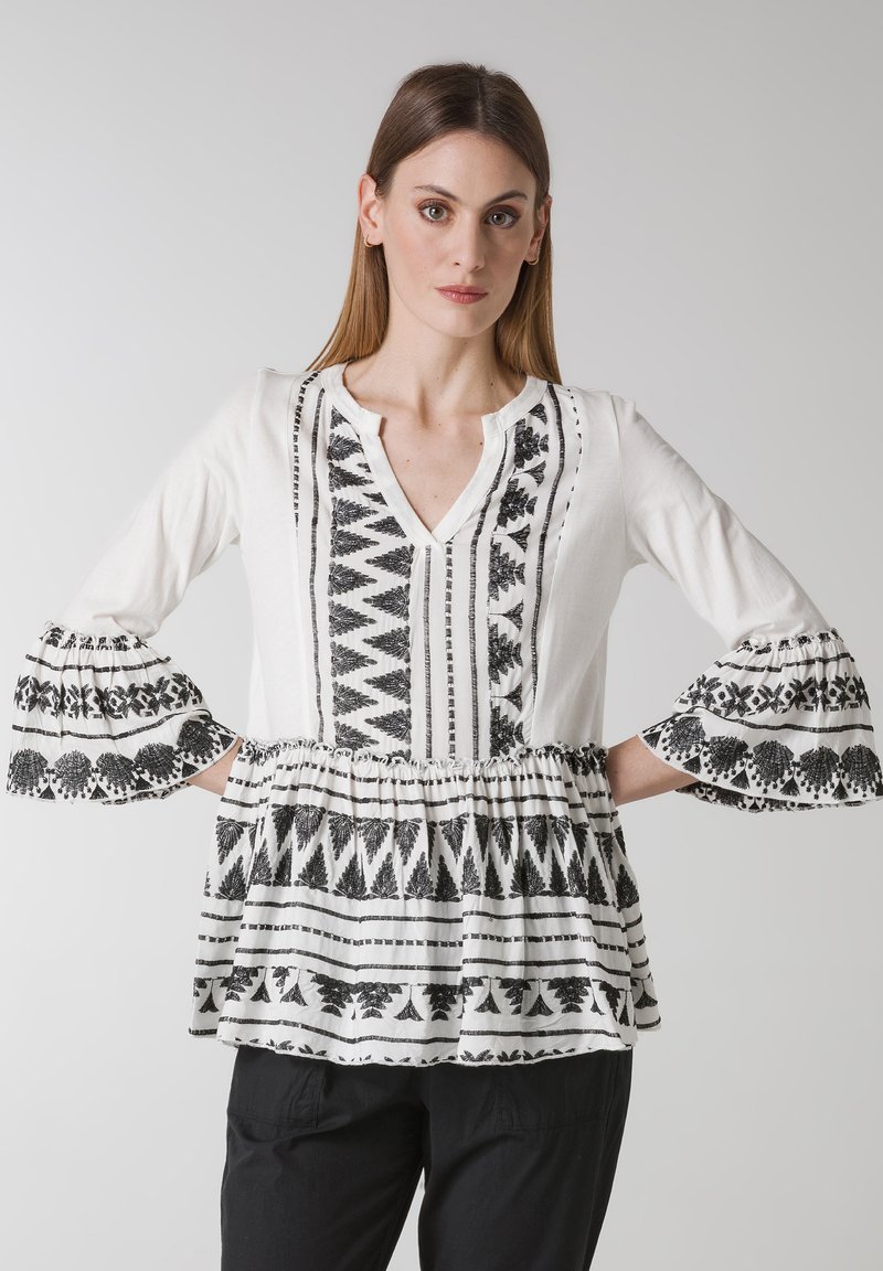 Vit blus med svart tribalbroderi, med V-ringning, utsvängda ärmar och en draperad midja. Mjuk, vävd tyg med en strukturerad mönster.