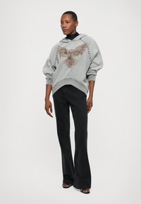 Light gray oversized hoodie featuring an eagle graphic, relaxed fit sleeves, and a ribbed hem, paired with flared black jeans.