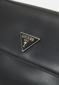 Cartera de cuero negro con superficie texturizada, herrajes plateados y un logotipo triangular que presenta 'GUESS USA' y un signo de interrogación.
