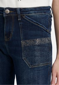 Close-up of dark blue jeans with metallic stud embellishments on the front pocket and a decorative button on the waistband.