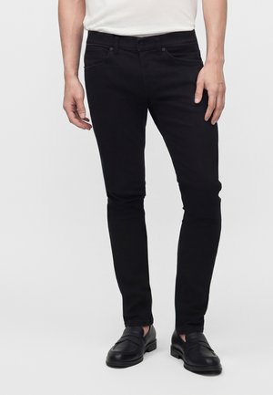 Mand iført slim-fit sorte bukser, sorte læder loafers og en hvid t-shirt, stående mod en ensartet lys baggrund.