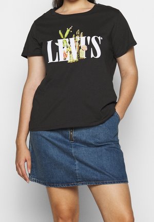 Persona vistiendo una camiseta negra de Levi's con diseño floral de cactus y una falda de mezclilla azul, con una mano en el bolsillo, de pie contra un fondo blanco.